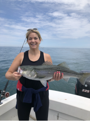 Woman hold large striped bass, standing in the stern of charter fishing boat