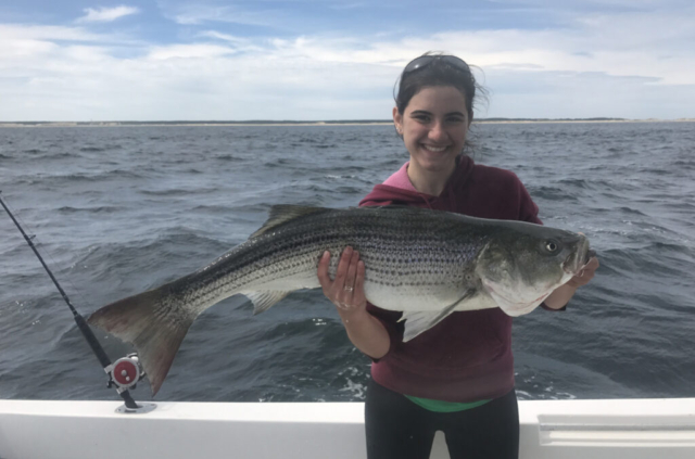 Smiling girl holding almost-four foot striped bass on deck of boat, ocean and blue sky in background